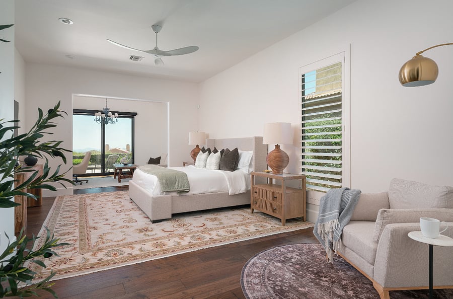 The primary suite is an oversized retreat spanning a king bed with Persian rug and seating area, a dedicated reading nook with oversized armchair and brass arc floor lamp, and a private workspace with full desk, sofa, and direct slider access to the patio with mountain views. Plantation shutters, ceiling fan, and a chandelier in the workspace alcove complete the suite.