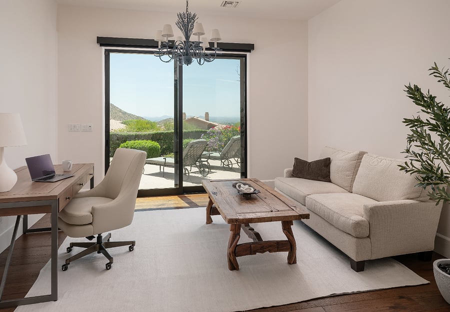 The primary suite is an oversized retreat spanning a king bed with Persian rug and seating area, a dedicated reading nook with oversized armchair and brass arc floor lamp, and a private workspace with full desk, sofa, and direct slider access to the patio with mountain views. Plantation shutters, ceiling fan, and a chandelier in the workspace alcove complete the suite.