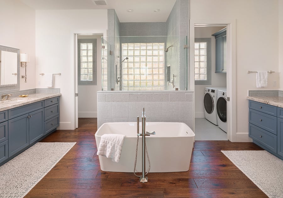 The primary ensuite includes a freestanding soaking tub, oversized glass-enclosed walk-in shower with bench and floor-to-ceiling tile, dual vanities with stone countertops and custom blue cabinetry, and a private laundry room with full-size LG washer and dryer and additional cabinetry.