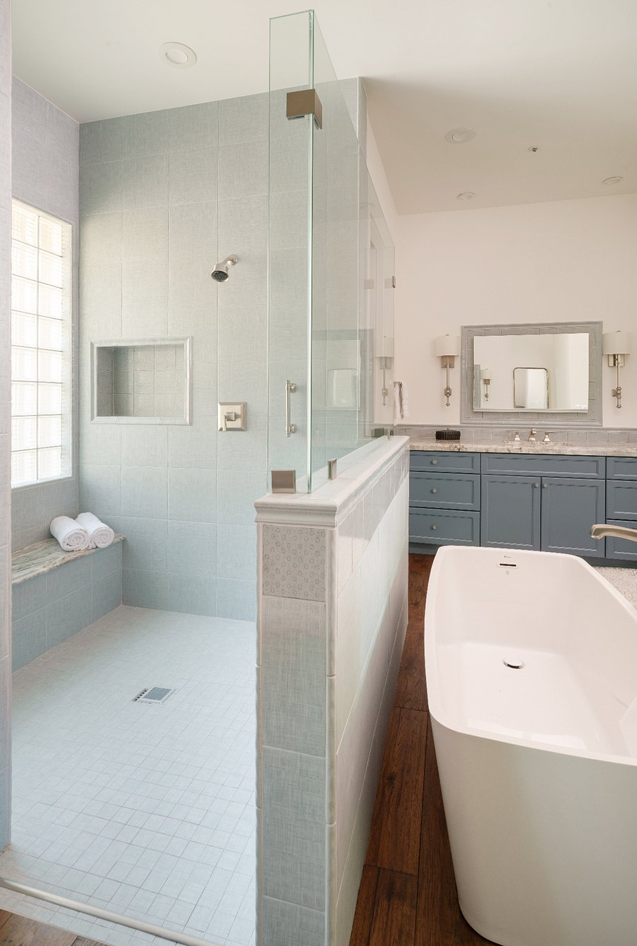 The primary ensuite includes a freestanding soaking tub, oversized glass-enclosed walk-in shower with bench and floor-to-ceiling tile, dual vanities with stone countertops and custom blue cabinetry, and a private laundry room with full-size LG washer and dryer and additional cabinetry.