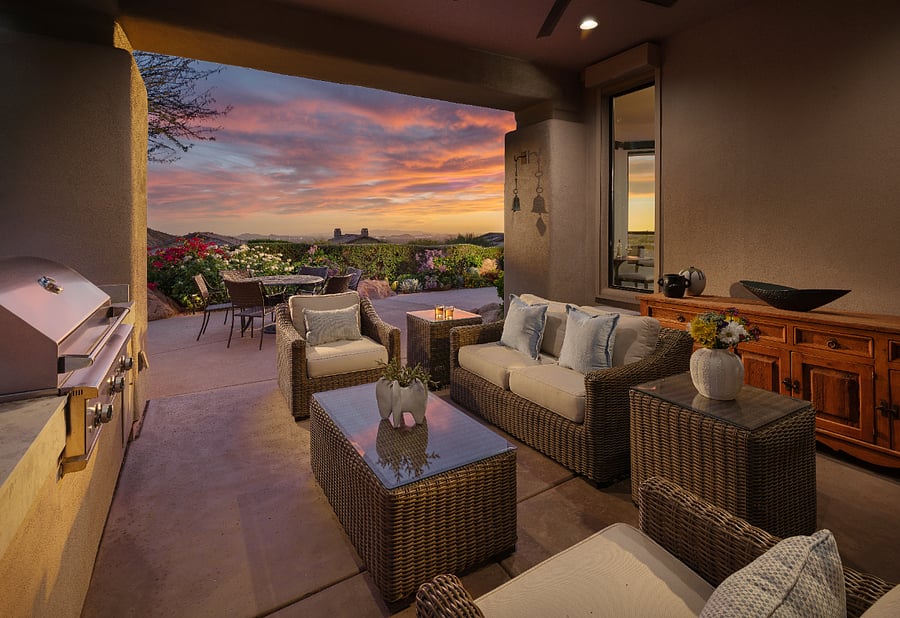 Covered outdoor living area with wicker sectional seating, built-in gas grill, and unobstructed views of Troon Mountain and city lights at sunset.